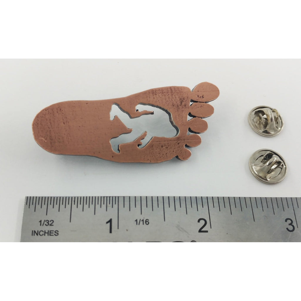 A handcrafted Sasquatch Bigfoot Footprint Pin next to a ruler, available for sale at Creativeartisan.com - the image offers a sense of scale